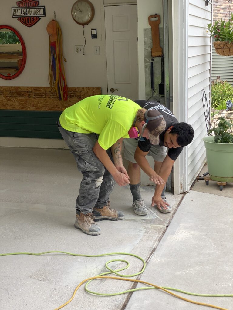 Two people work on garage floor leveling. Tools, a Harley-Davidson sign, and garden plants are visible. Casual and practical attire worn.