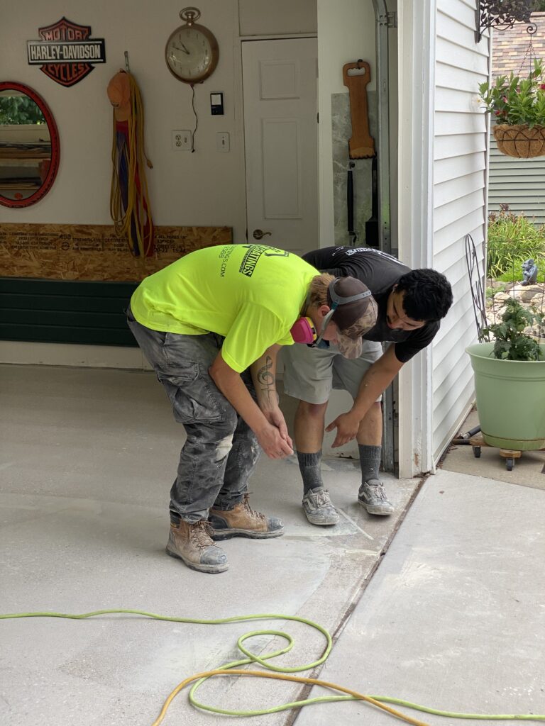 Two persons working on a garage floor, one wearing a bright shirt. Harley-Davidson sign and clock visible on the wall.