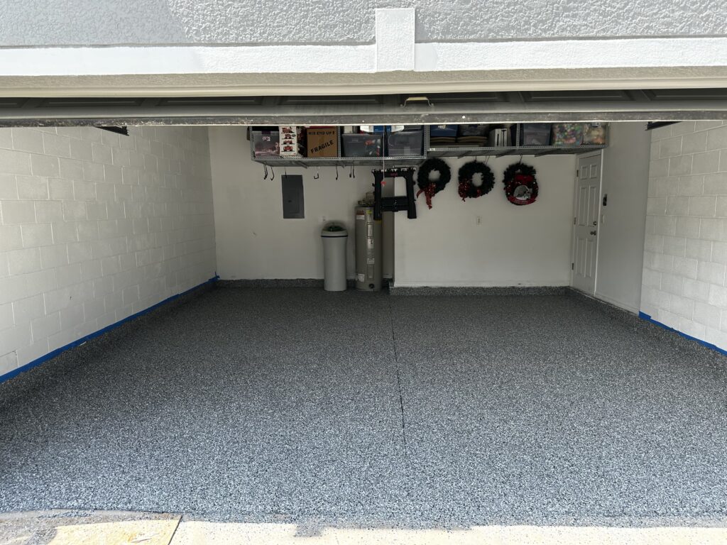 Empty garage with speckled gray floor, white walls, and holiday wreaths on the back wall. Shelves hold boxes and miscellaneous items above.