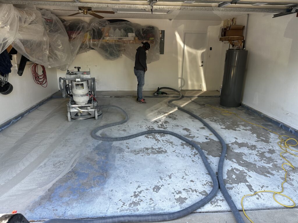 A person with equipment works on a garage floor. There are tools hanging, and the space has a partially stripped, uneven surface.