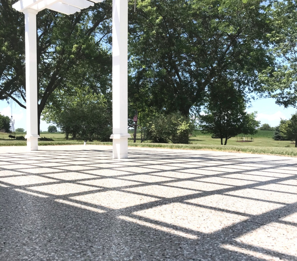 A white pergola casts geometric shadows on a concrete patio, surrounded by lush green trees under a clear blue sky.