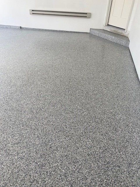 A clean, gray speckled epoxy floor in a garage space, terminates at a white door and gray baseboard heater on the wall.